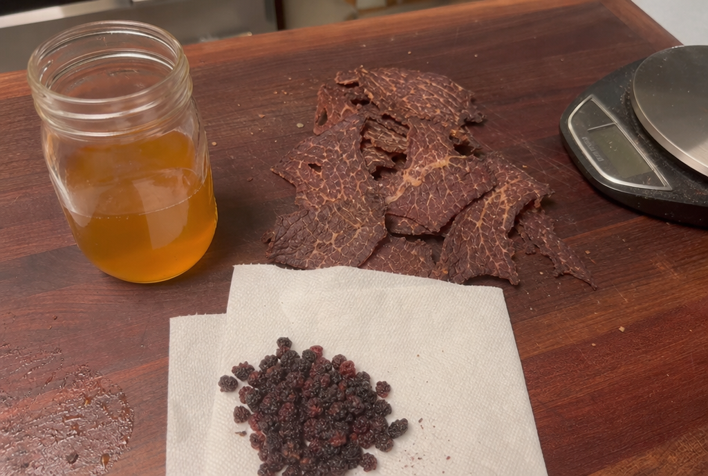 Homemade pemmican ingredients on a wooden board: dried beef jerky, a mason jar of liquid rendered tallow, and wrinkled dried berries before mixing