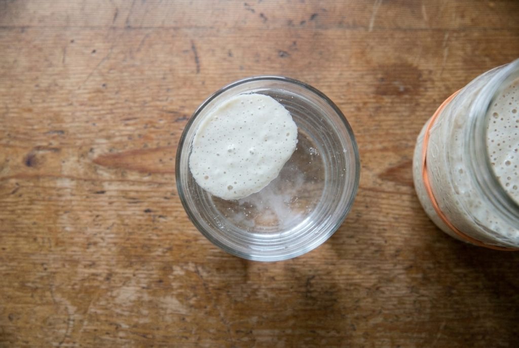 Overhead view of a flat oval blob of sourdough starter floating in a clear glass of water during the float test, on a weathered wood table.